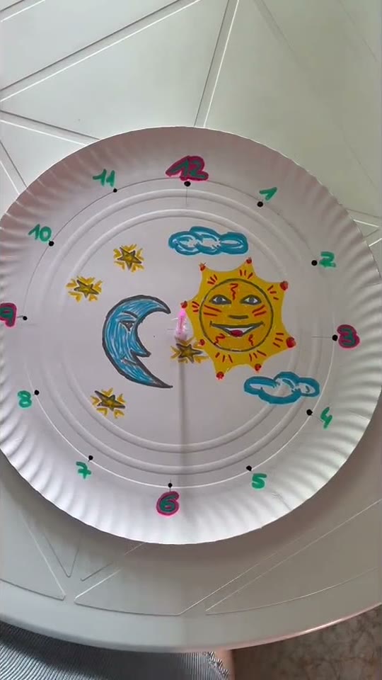 Rellotge de sol fet amb una base circular de cartró decorada a mà amb un sol somrient, una lluna blava i núvols, amb les hores marcades al voltant